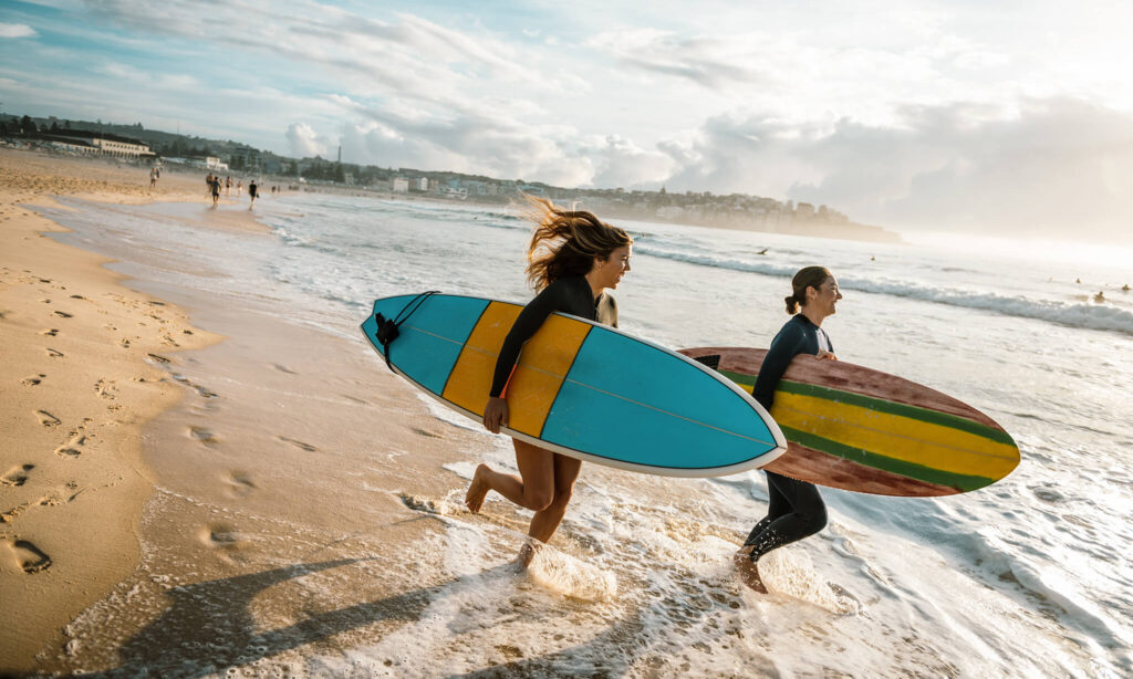Dos mujeres jóvenes corren hacia el oleaje de la costa norte con tablas de surf.