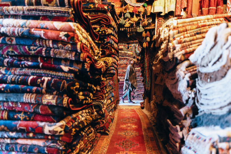Una mujer hojea pilas de alfombras finas en un mercado.