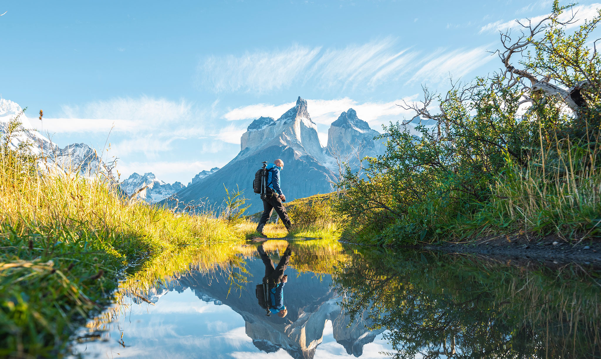 Un excursionista con equipo fotográfico cruza un estanque mientras a lo lejos se alzan afilados picos montañosos.