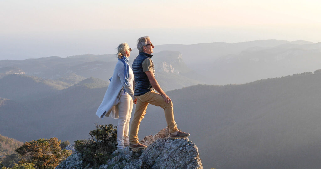 Una pareja de personas mayores sobre una roca con vistas a unas montañas lejanas.